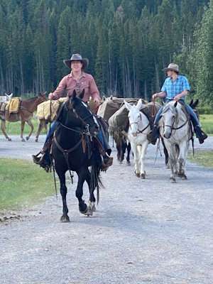 Two cowboys wearing cowboy hats and riding horses with pack donkeys in tow