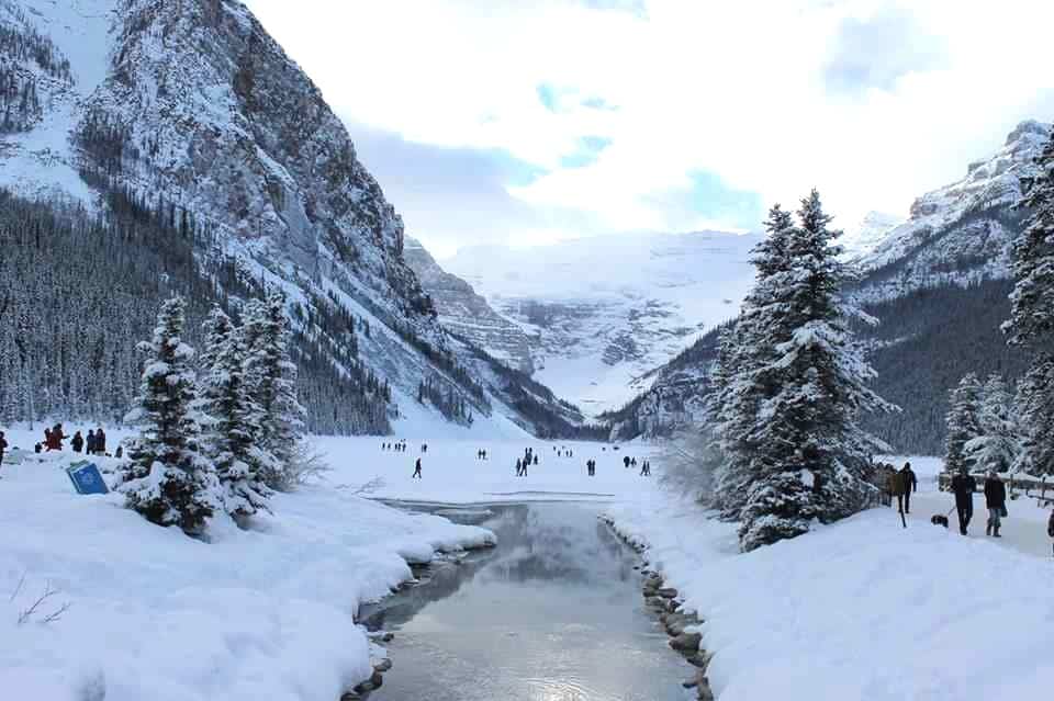 Lake Louise in winter with water, and the frozen lake and mountains in the background. People are ice skating on the lake.