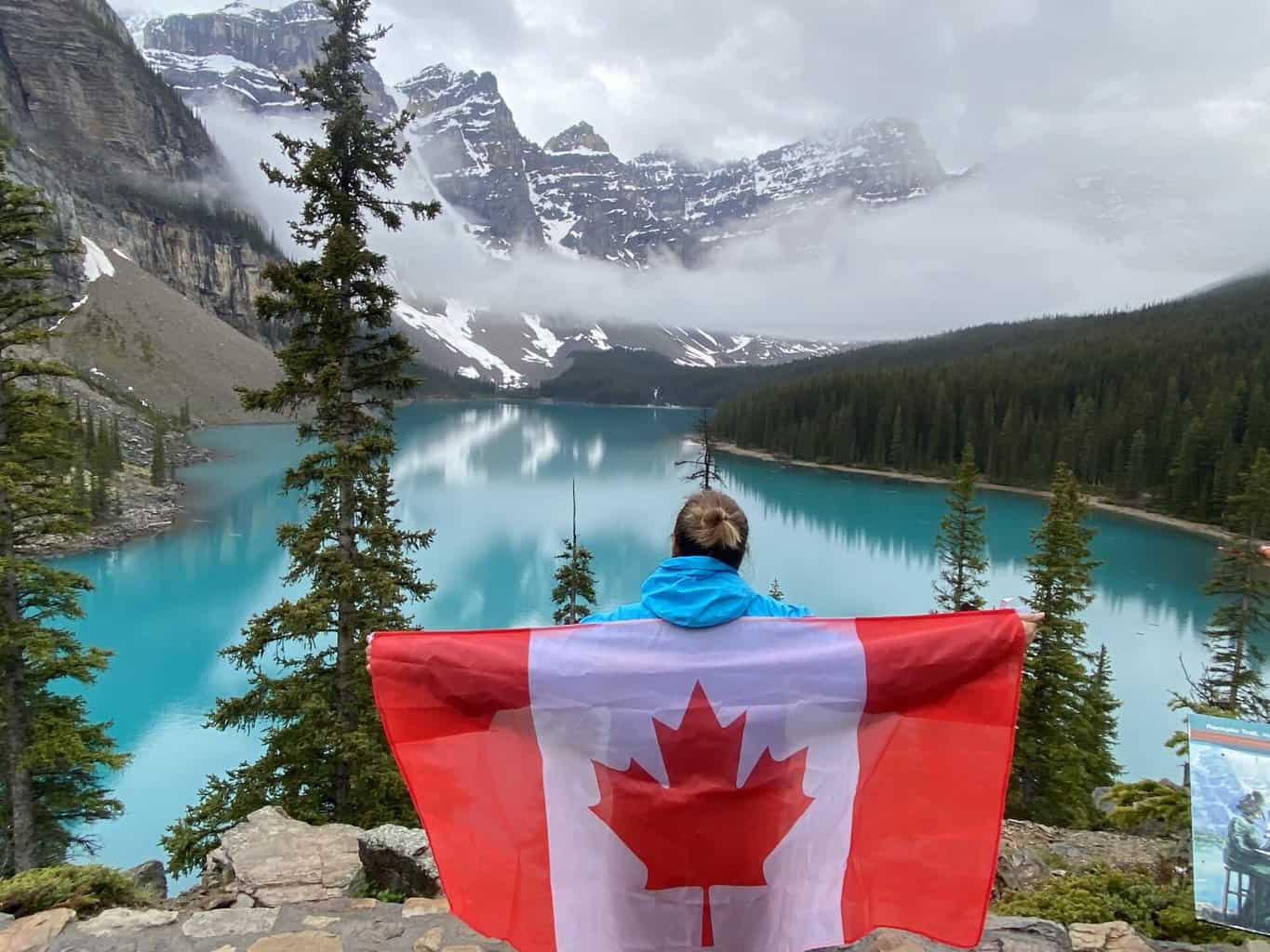 Woman with Canada flag standing in front of turquoise Moraine Lake, pine trees, and mountains