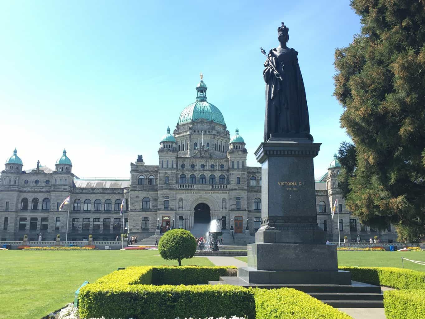 Victoria legislature building and grassy grounds with statue of Queen in front of brick castle like building with green turrets