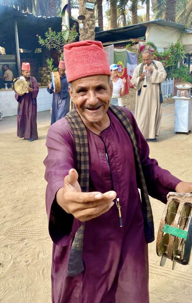An Egyptian mad with a red hat and purple caftan holding out his hand
